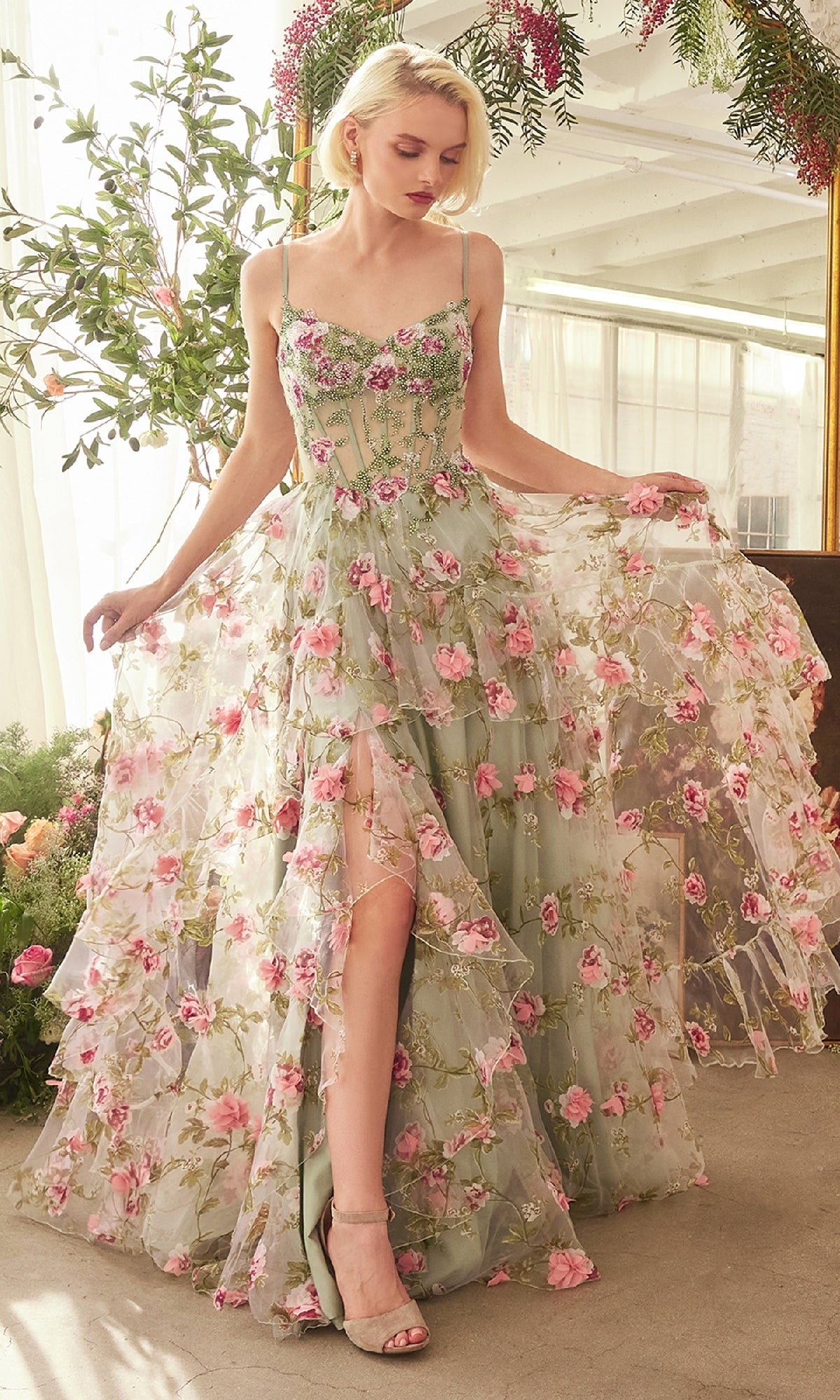 A woman stands indoors near plants, holding out the sides of her Andrea & Leo A1420 Sage Long Floral Prom Dress featuring a sheer corset and pink floral patterns.